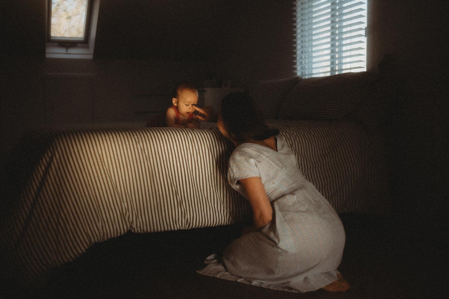 baby laying down in bed while mom touches his nose while she's seated on the floor, captured during an in-home motherhood session in Dorval, Montreal