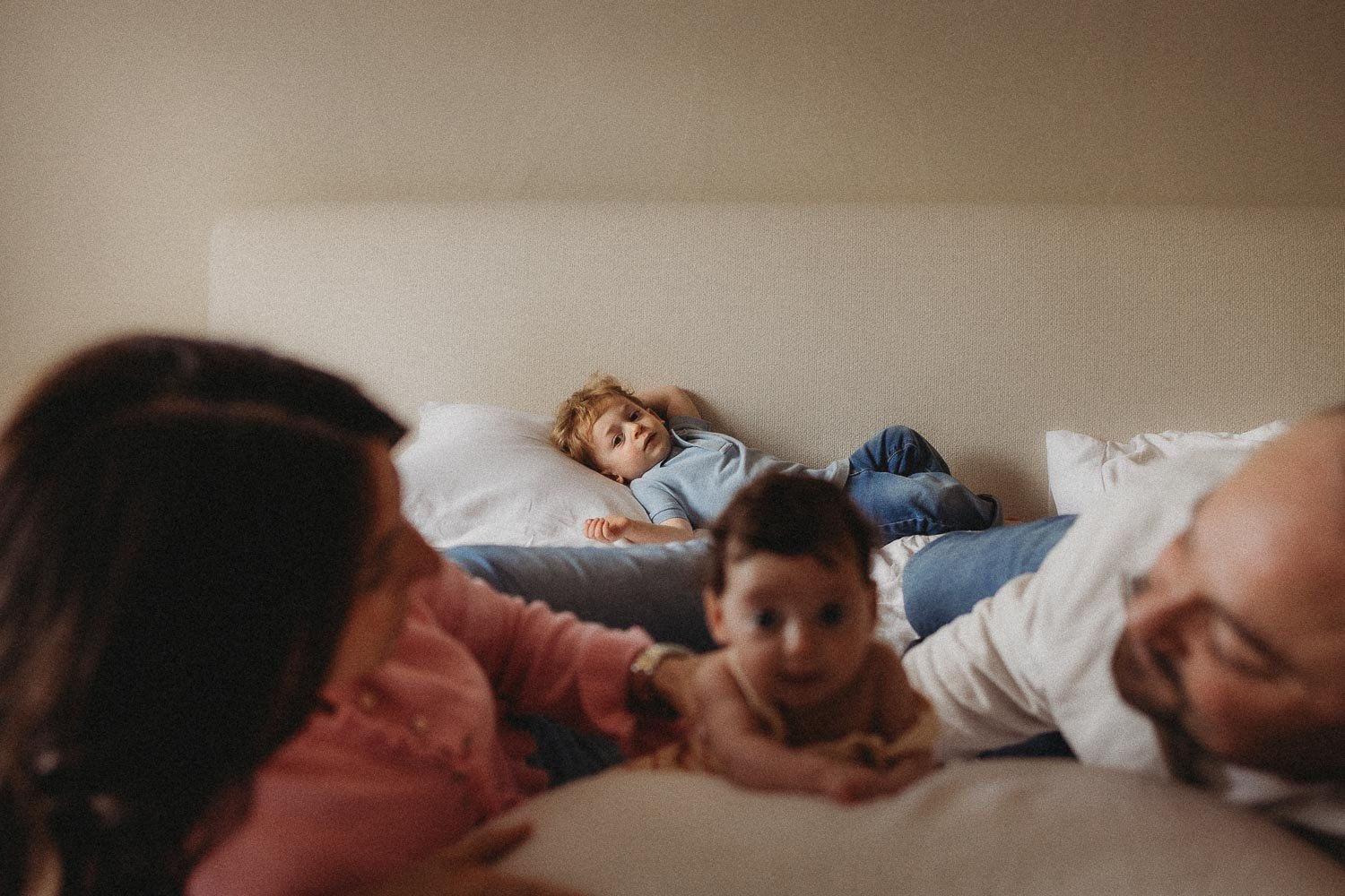 A family of four laying down in bed during an in-home newborn photoshoot in Hampstead, Montreal. The mom, dad and baby are out of focus and in the foreground, the boy is resting on a pillow, looking at the camera and in focus