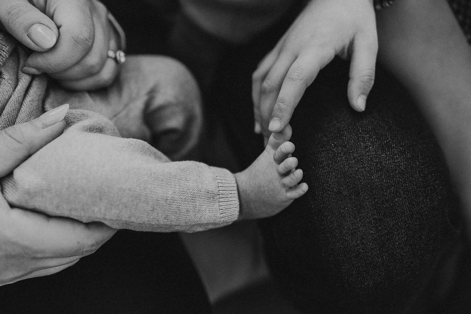 Black and white photograph of a baby's hand being gently held by an adult's hand, with another adult's hand nearby, resting on a person's knee.
