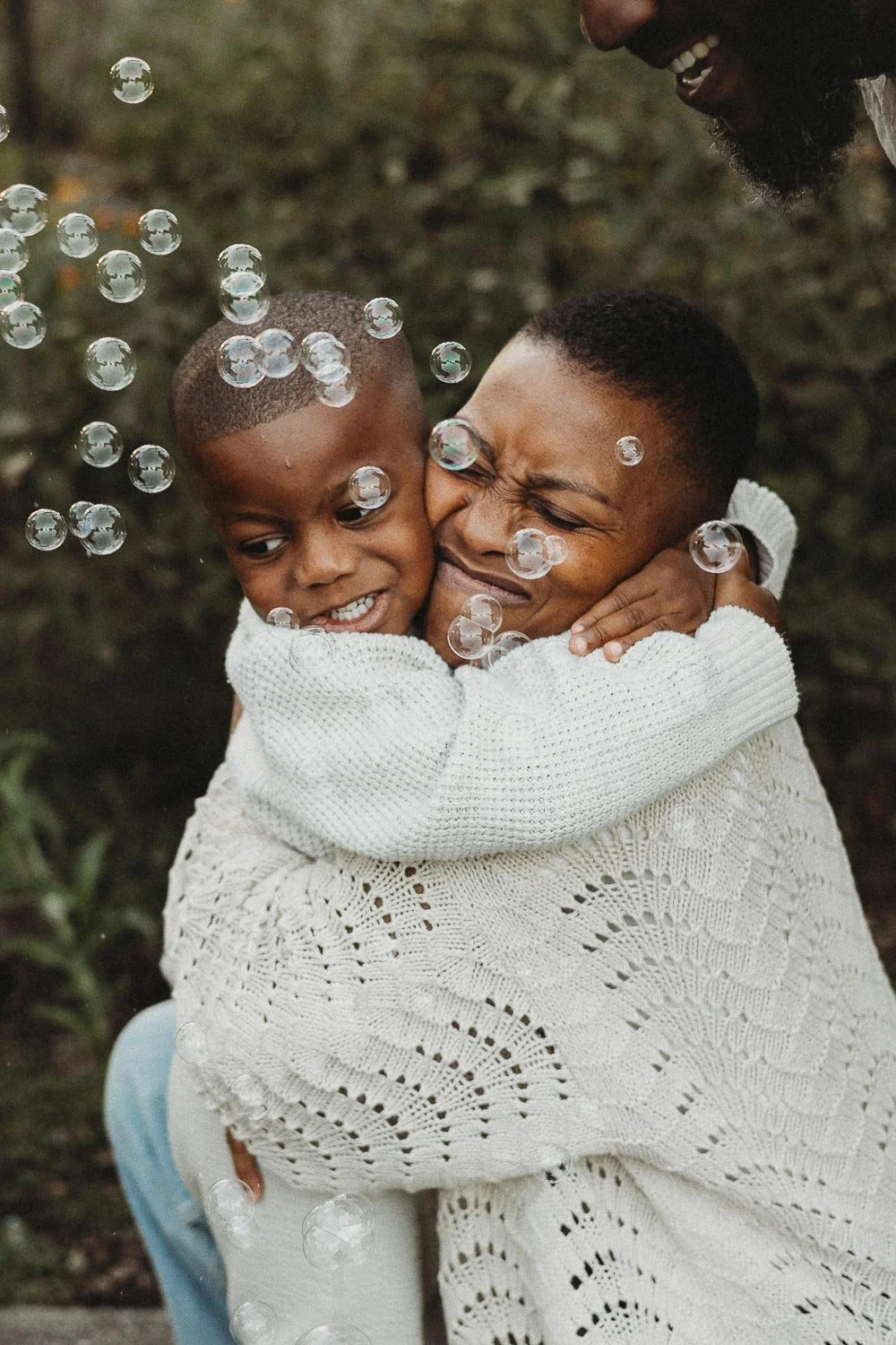 Outdoor Family photo session of a mom of dark skin colour, hugging tightly her son, while bubbles fly in front of their faces at the Montreal Botanical Garden