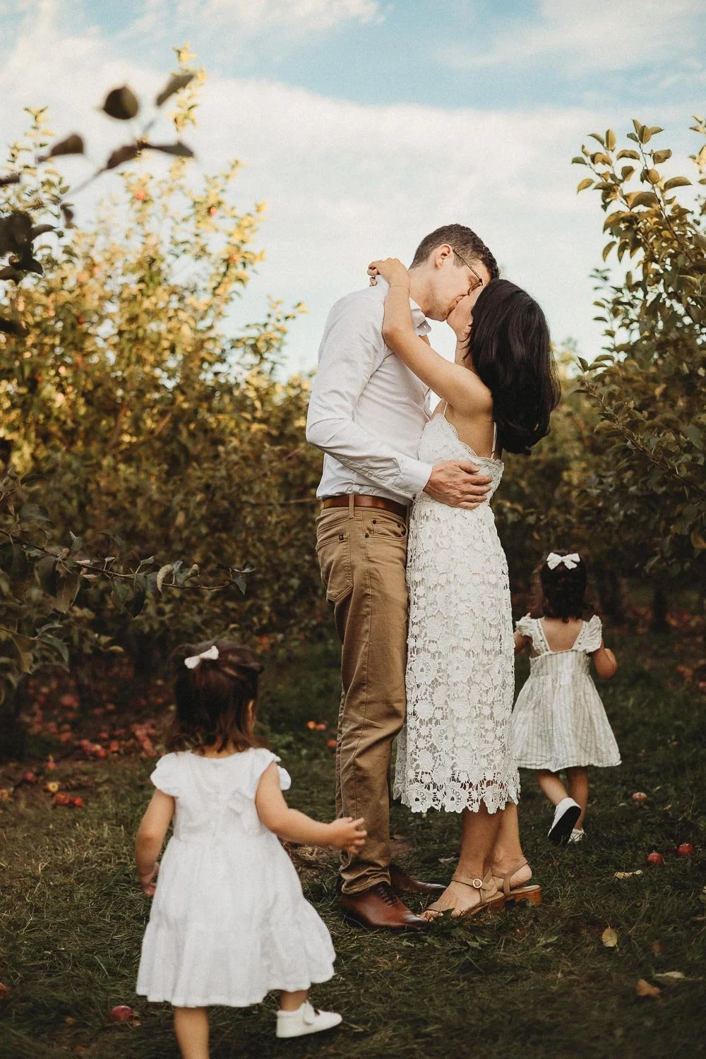 A couple shares a kiss in an orchard with two young girls nearby, one walking away and the other reaching out captured at an outdoor Family Photoshoot near Montreal.