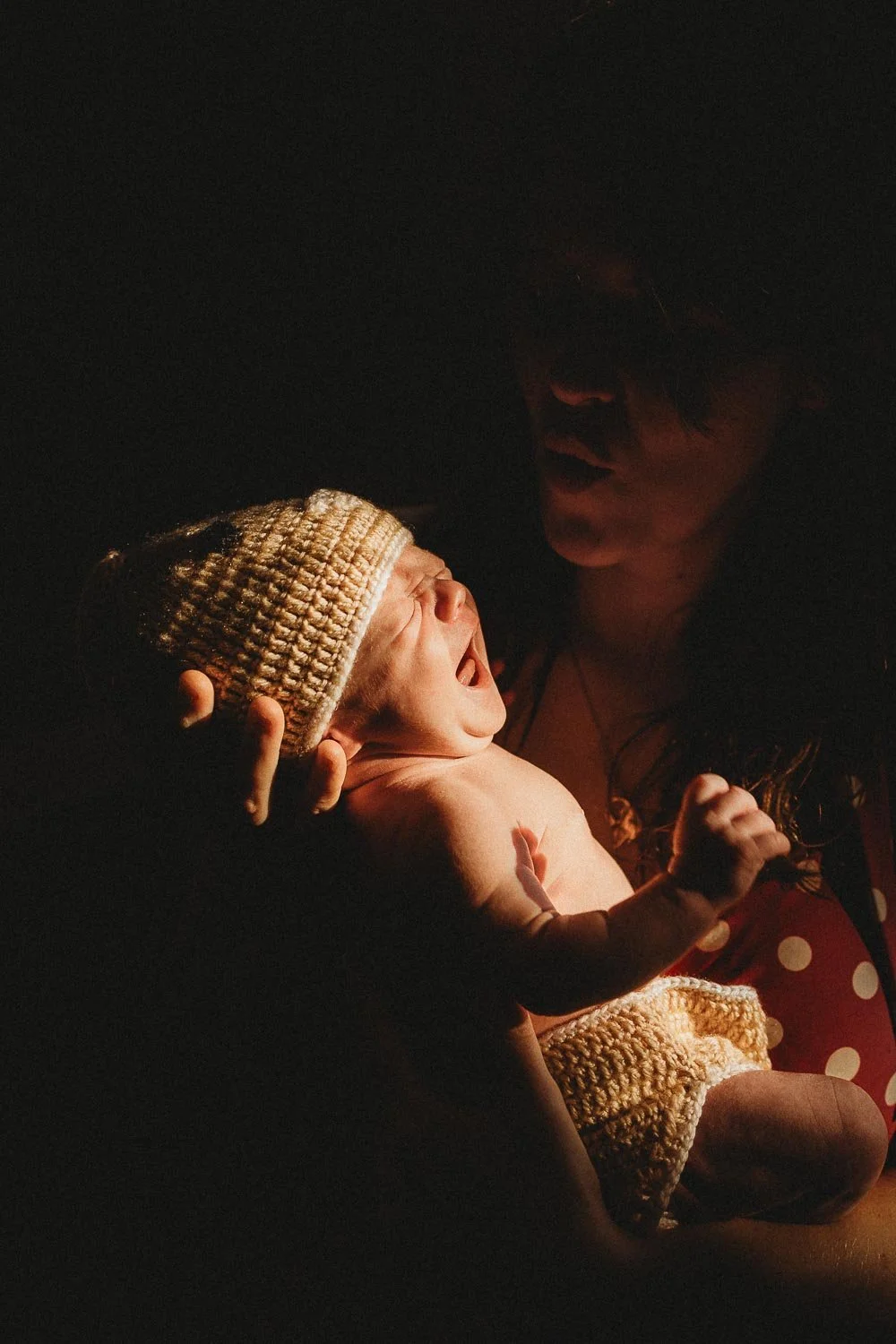 Newborn baby being held by his mom. The room is dark and the baby is lit by natural window light. The mom is in the darkness and talking to baby