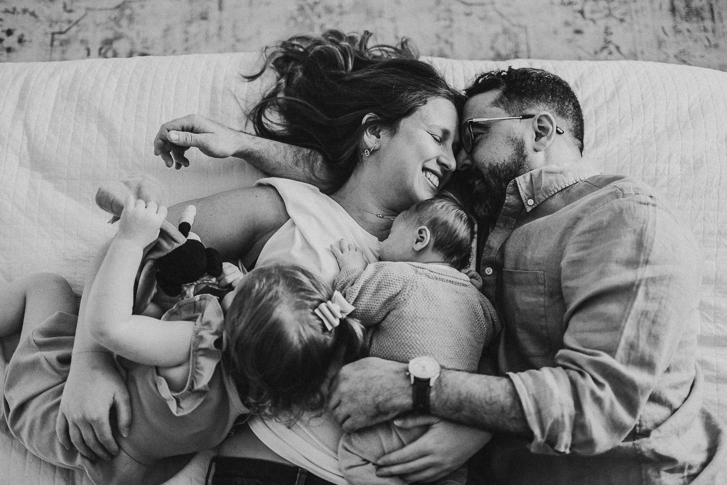 A family of four laying down in bed while a newborn photoshoot. The mom is holding the newborn baby on her chest, the dad is forehead to forehead with mom and holding them, and the toddler girl is resting her head on mom's belly. Mom is smiling