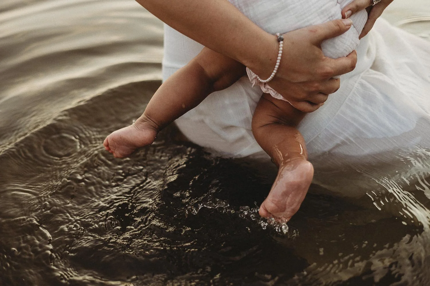 An outdoor motherhood photoshoot at Cap-Saint-Jacques portraying a mom holding her baby, both in white clothes, in shallow water, with the baby's legs and feet submerged and the adult supporting the baby from behind.