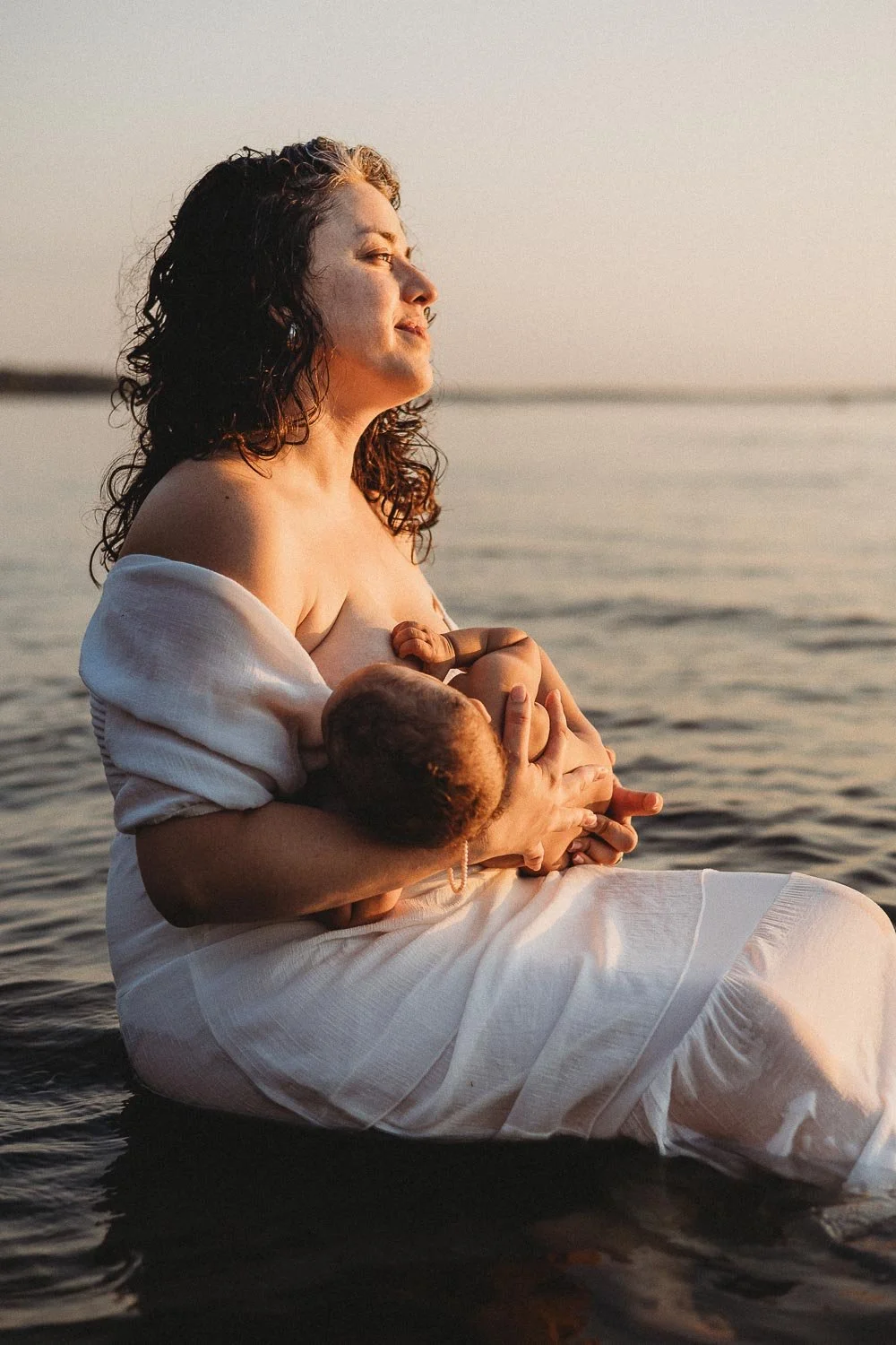 Mother breastfeeding her baby while looking at the sunset during a motherhood beach photoshoot at Cap-Saint-Jacques, Montreal