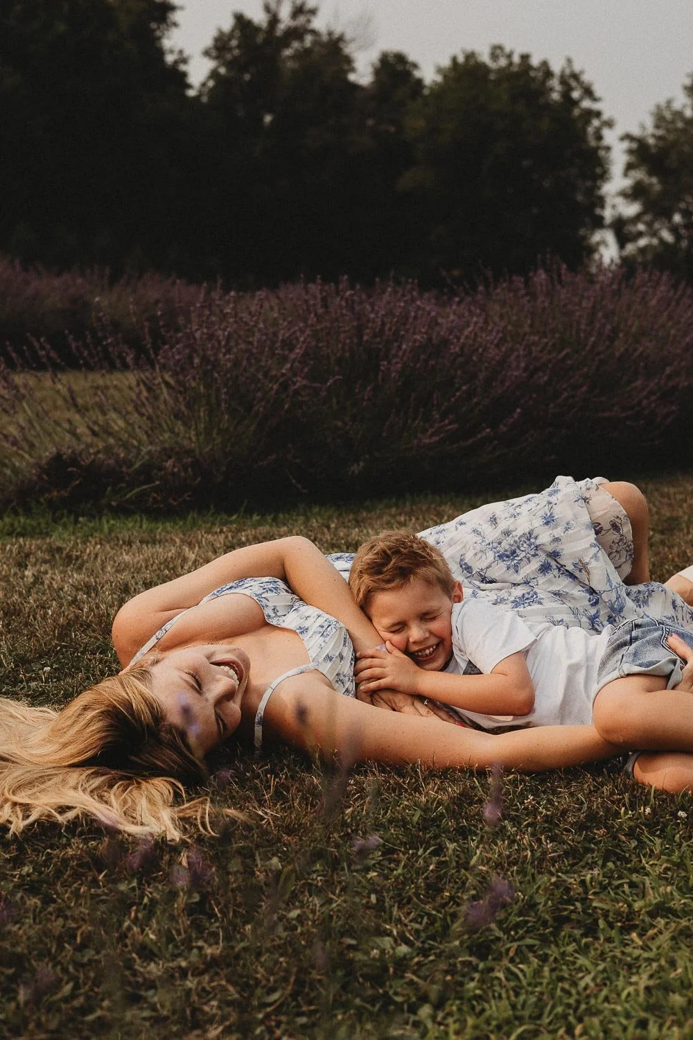 A woman and a young boy are lying on the grass outdoors, laughing and hugging each other at sunset, with purple flowering bushes and trees in the background enjoying an outdoor family photo session at La Maison Lavande, Quebec