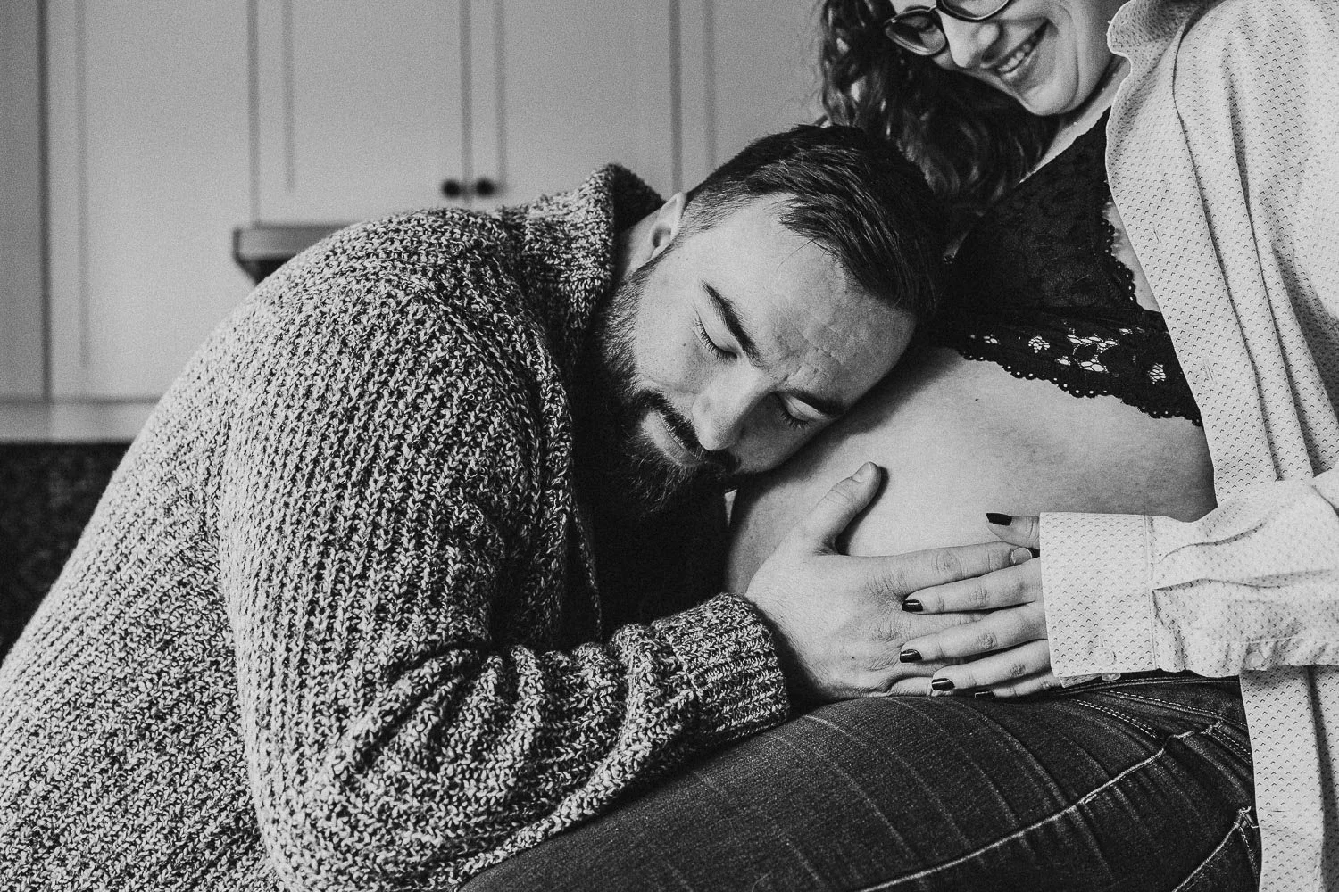 Pregnant woman smiling at husband who is resting his head on her pregnant belly while holding hands, captured during an in-home maternity photo session in Montreal