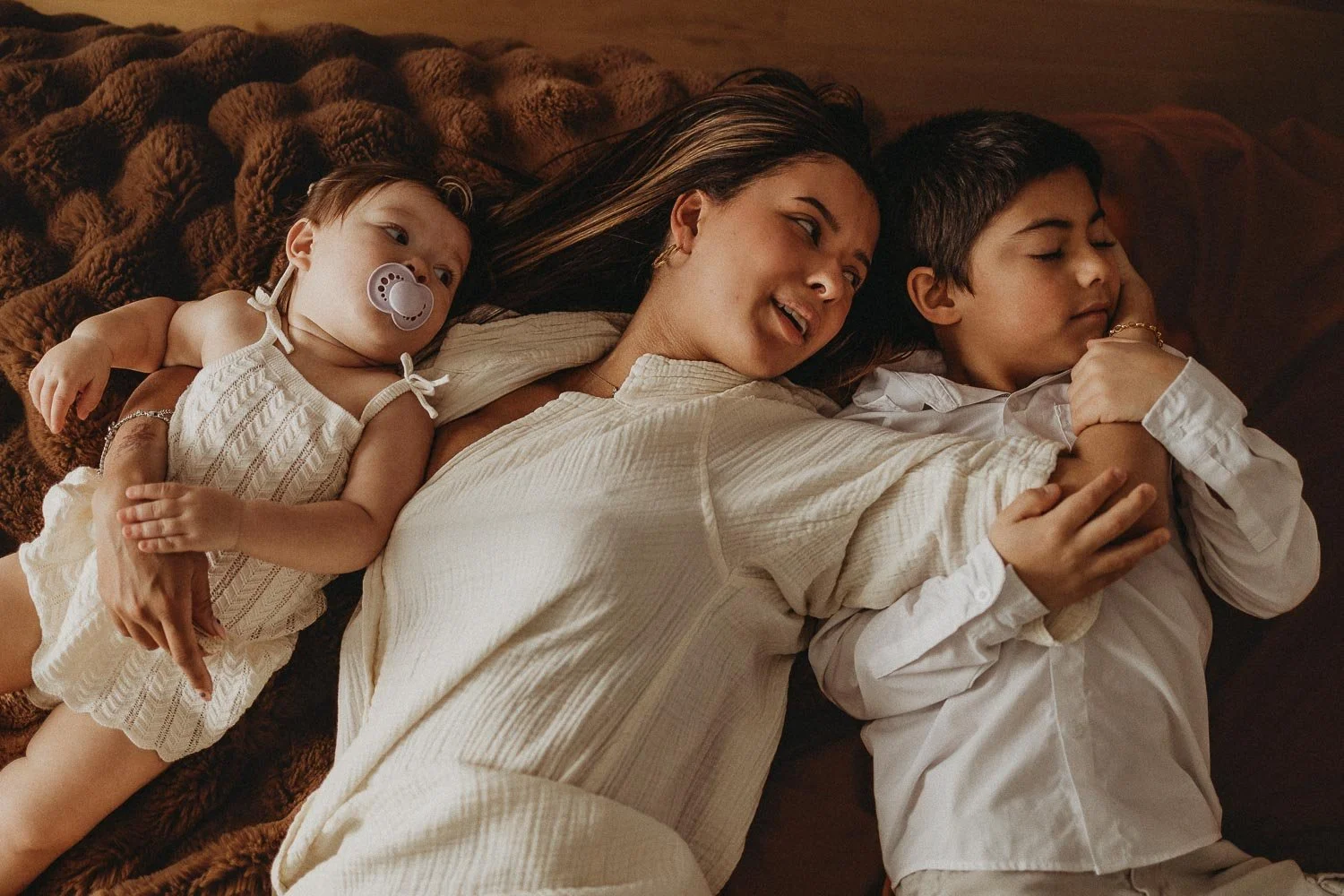 An in-home motherhood photo session in Montreal portraying a woman lying on a bed with two children, one a toddler with a pacifier, all smiling and cuddling.