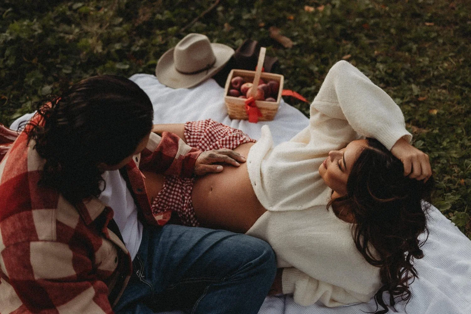 Pregnant woman laying down on a picnic blanket holding her hair while her male partner is resting his hand on her belly, captured during an outdoor maternity photoshoot in the Verger du Flanc Nord, Mont Saint-Hilaire