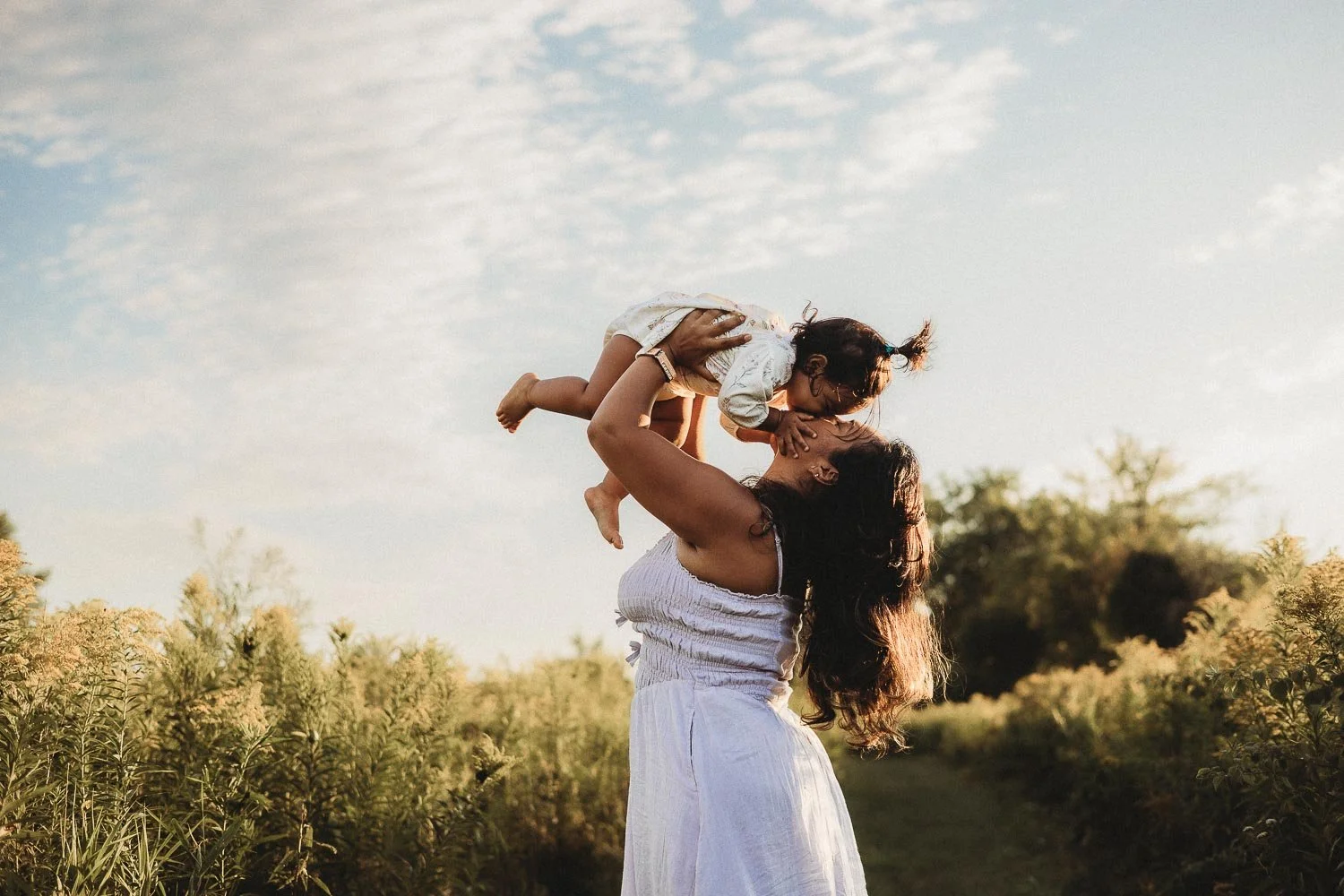 Mom raising her kid while kissing her on the face, at golden hour and surrounded by nature. Captured during an outdoor Motherhood photoshoot in Boucherville Island, Montreal