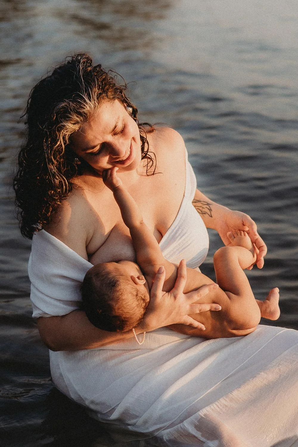 A mom breastfeeding her baby who is touching her face. Mom is smiling and gently touching baby's foot during a beach Motherhood Photoshoot at Cap-Saint-Jacques, Montreal