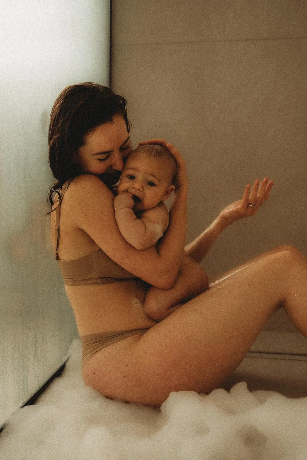 An in-home motherhood photoshoot where a mom is sitting down in a shower floor while holding her baby and smiling at him while touching the water that falls from the shower, in Dorval Quebec