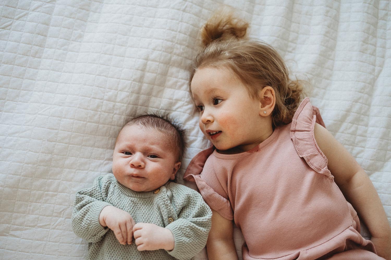 Newborn baby laying down in bed and looking at the camera whith sister laying down next to him, captured during an in-home newborn photoshoot in TMR, Montreal