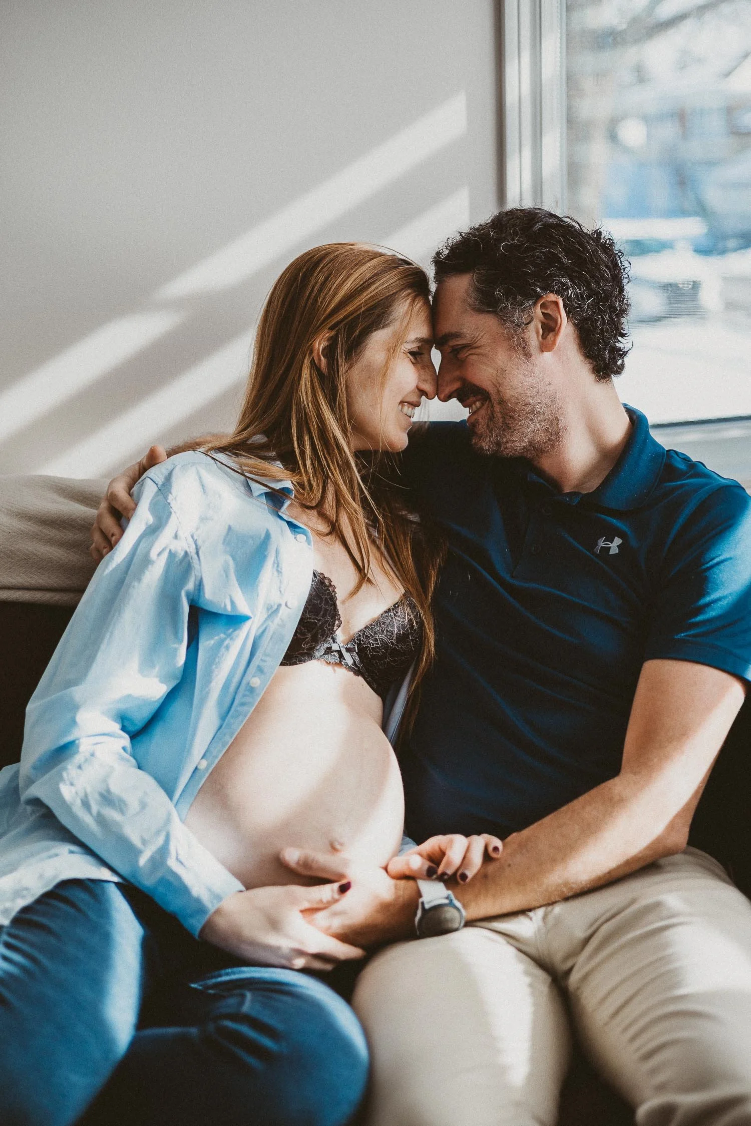 A couple, with the woman visibly pregnant, touching foreheads and smiling at each other inside a room with sunlight coming through a window.