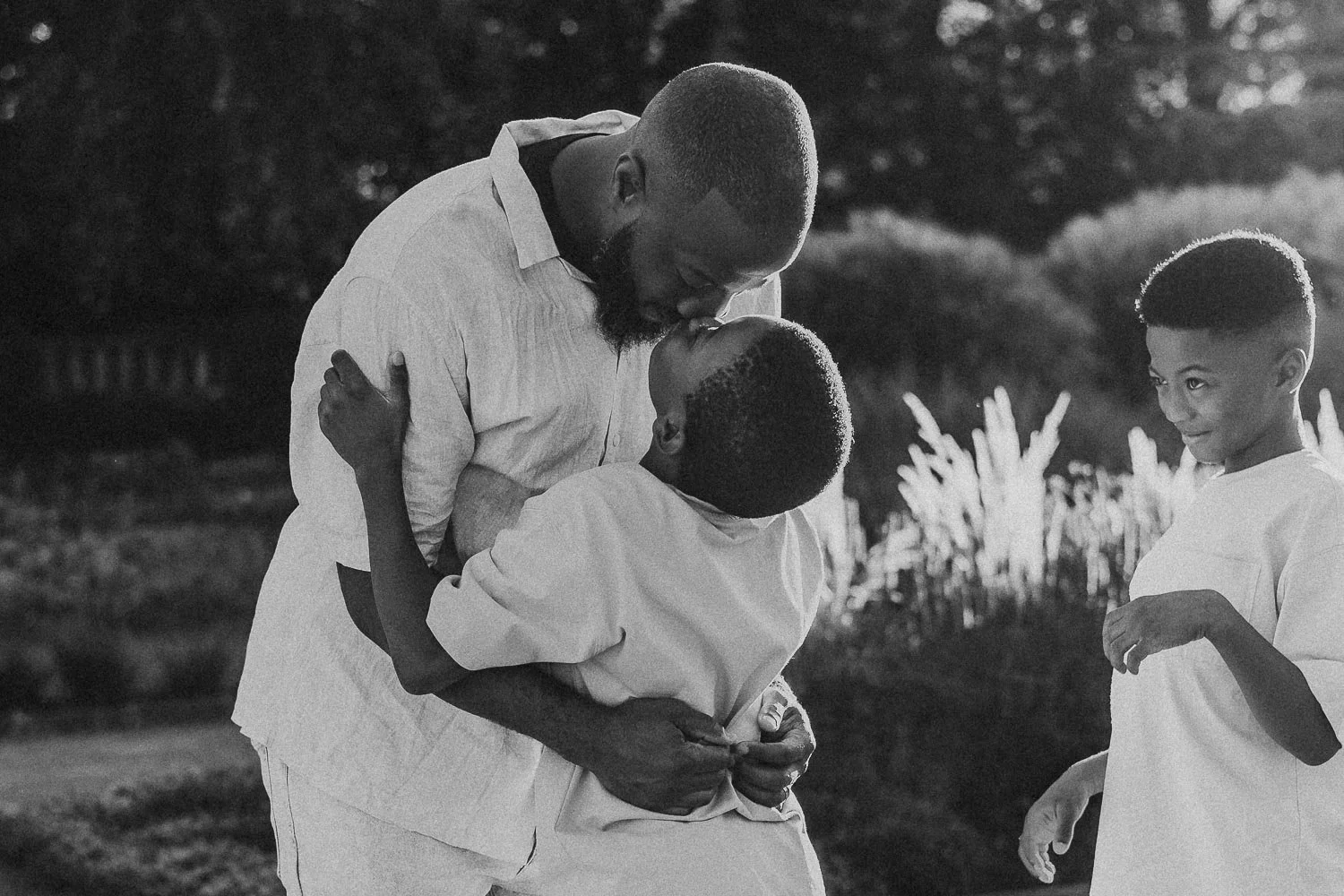 A man and boy sharing a kiss outdoors, with a girl standing nearby watching with a smile captured at the Montreal Botanical Garden during a family photoshoot