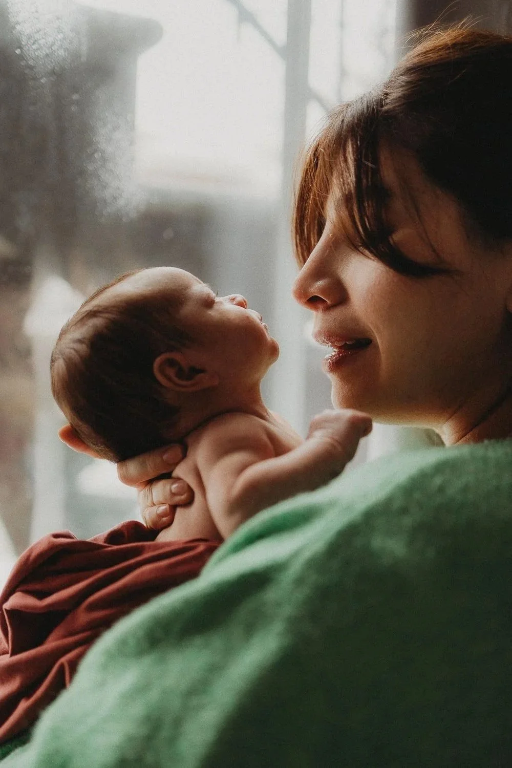 A mom holding her newborn baby and looking gently at him, close to a window and captured during an in-home newborn photo session at La Salle, Montreal