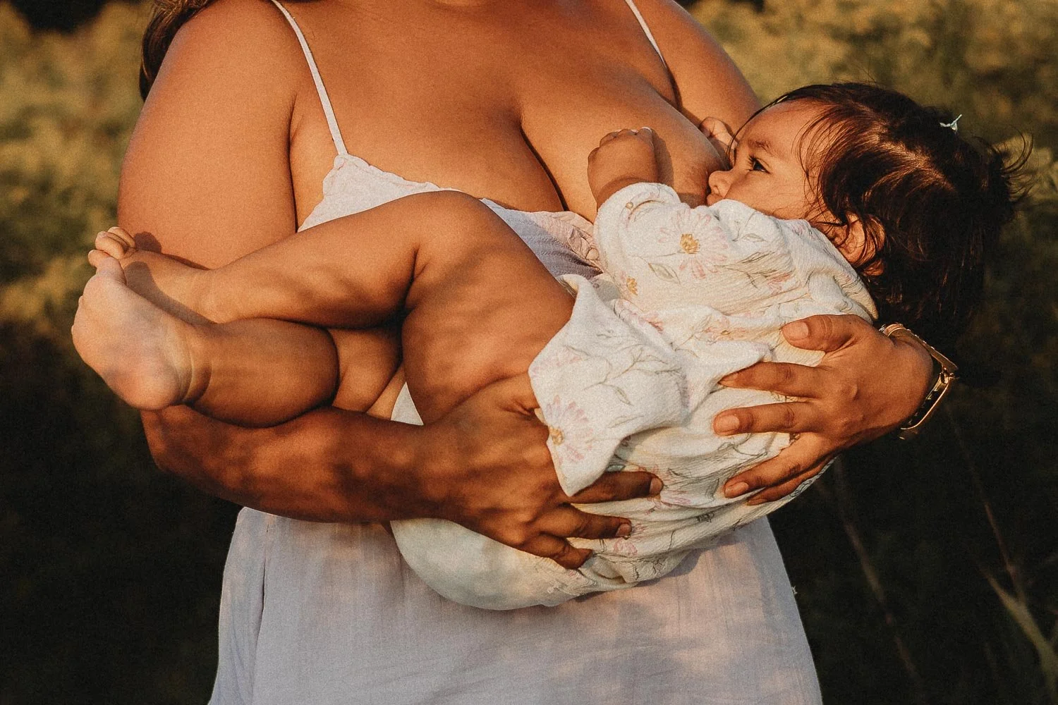 A closed-up photo of a mom holding her baby girl with both hands while she's breastfeeding, some shadows of the trees are on them creating an artistic image, captured during an outdoor motherhood photoshoot at the Boucherville Island, in Montreal