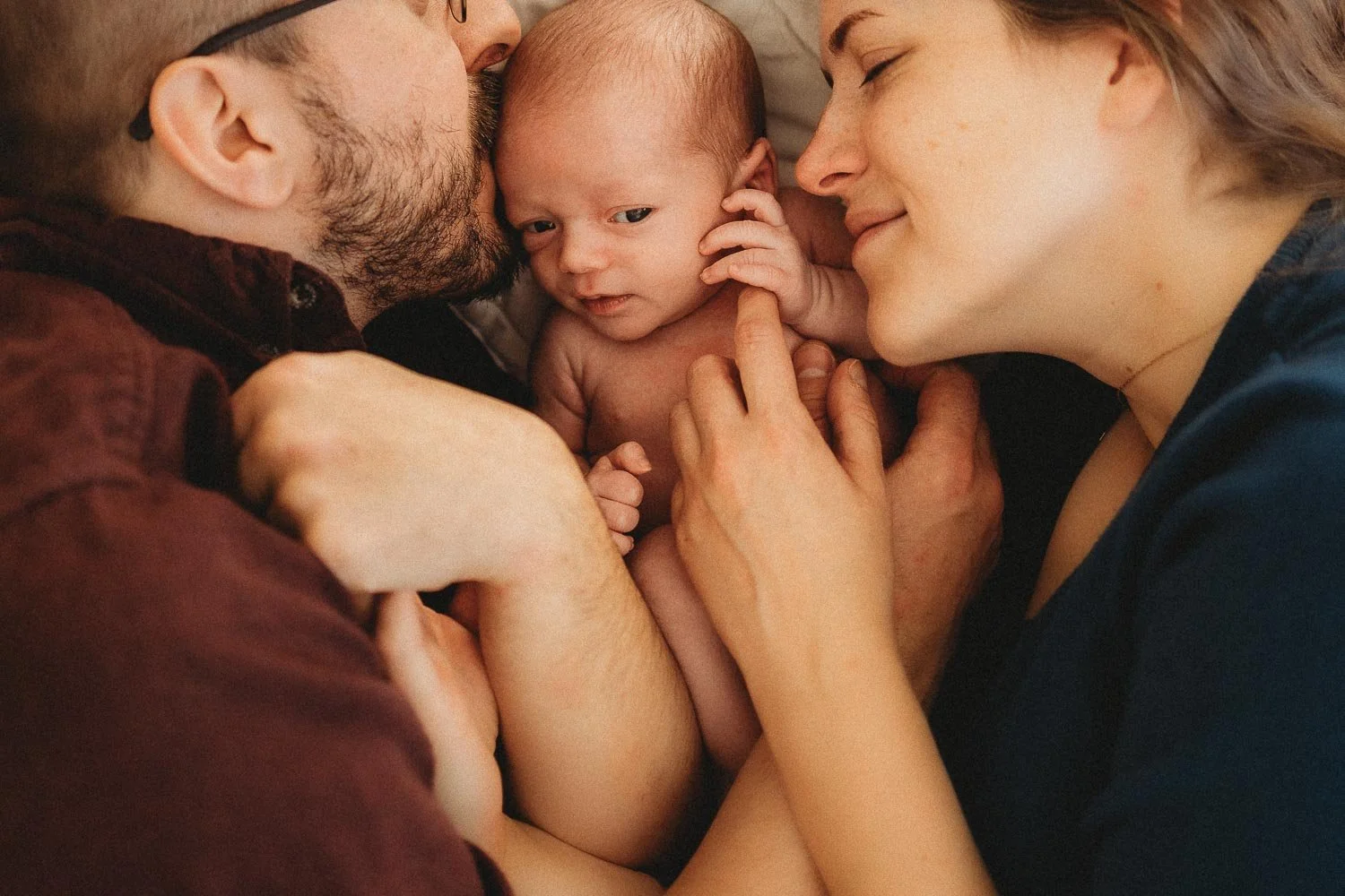 Mom, dad and newborn baby laying down in bed, cuddling and holding hands captured during an in-home photo session in Montreal