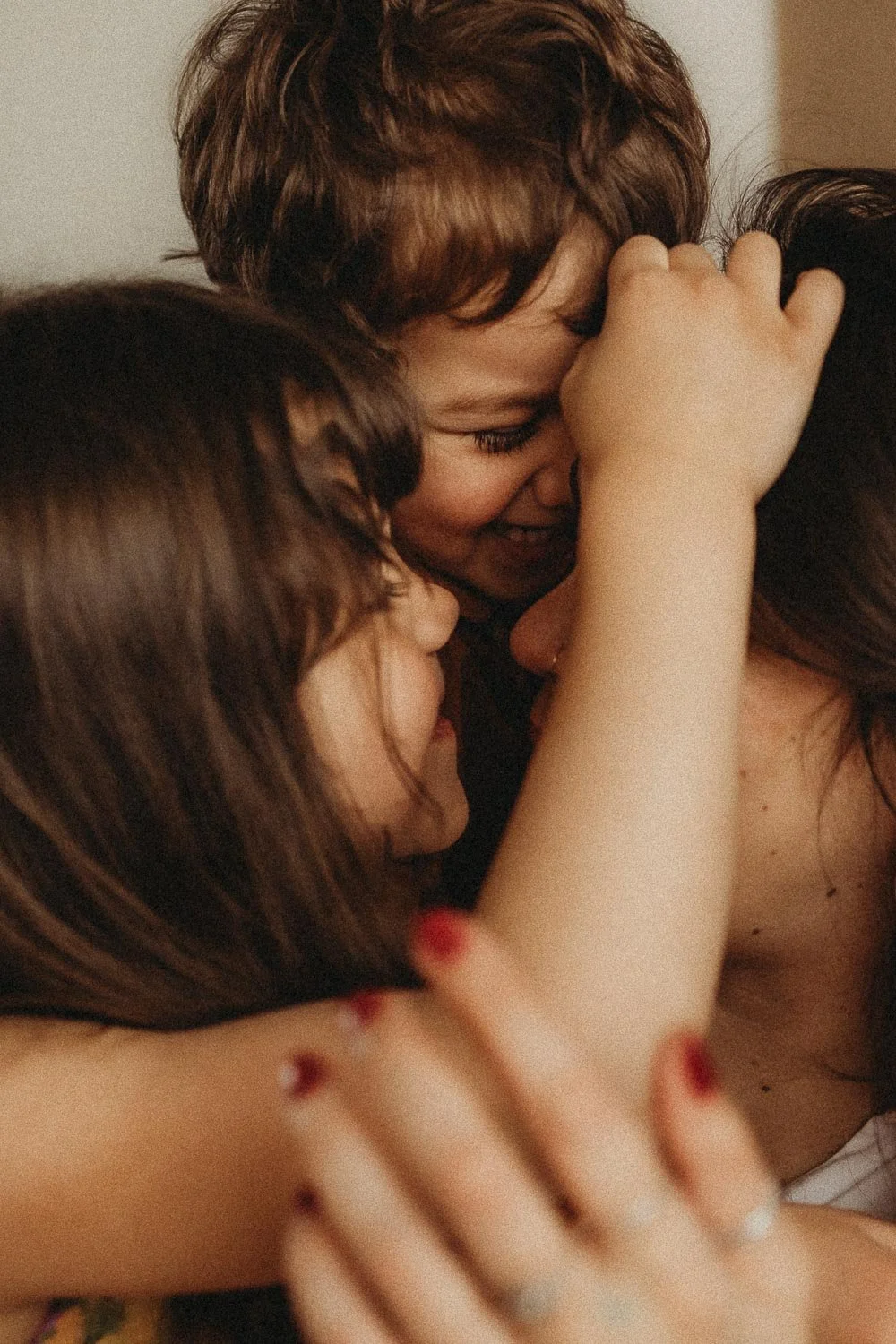 A woman happily hugs a young child indoors, with a decorative wicker background and a potted plant visible during a motherhood photoshoot in Montreal.