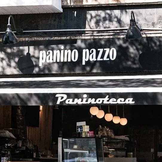 Storefront with black awning displaying white text 'panino pazzo' and 'Paninoteca'; outdoor lighting and interior visible through open entrance.