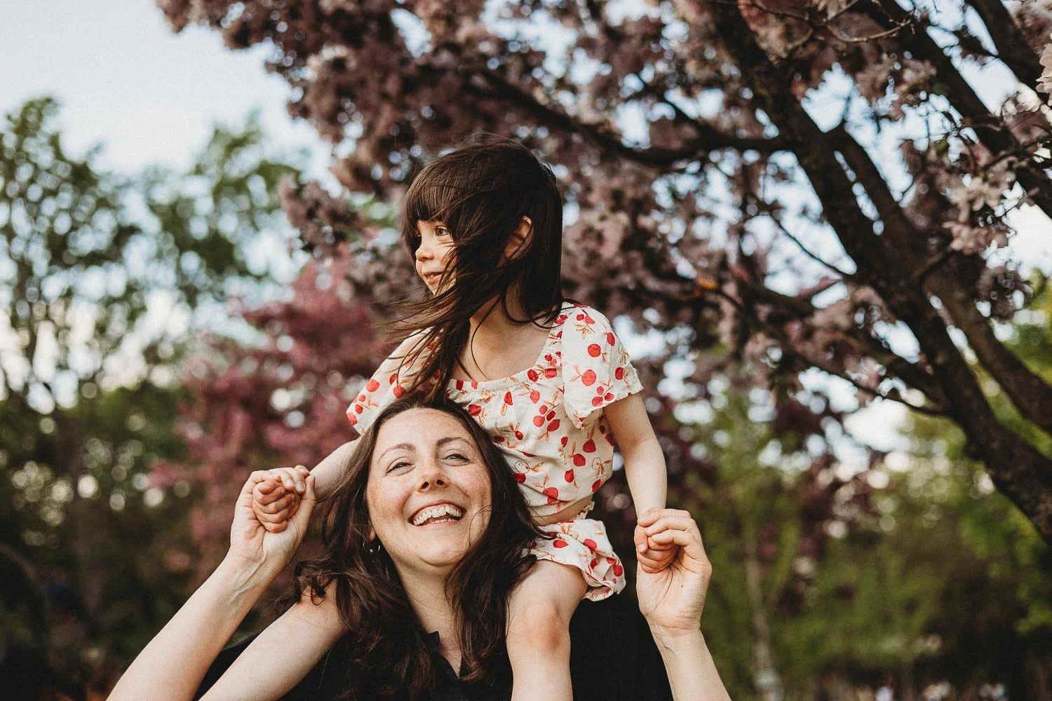 Mom carrying her daughter on her shoulders while holding her hands gently, mom sliming and girl looking happily looking away, cherry blossom trees are at the background, captured at Westmount Park, Montreal during an outdoor family photoshoot