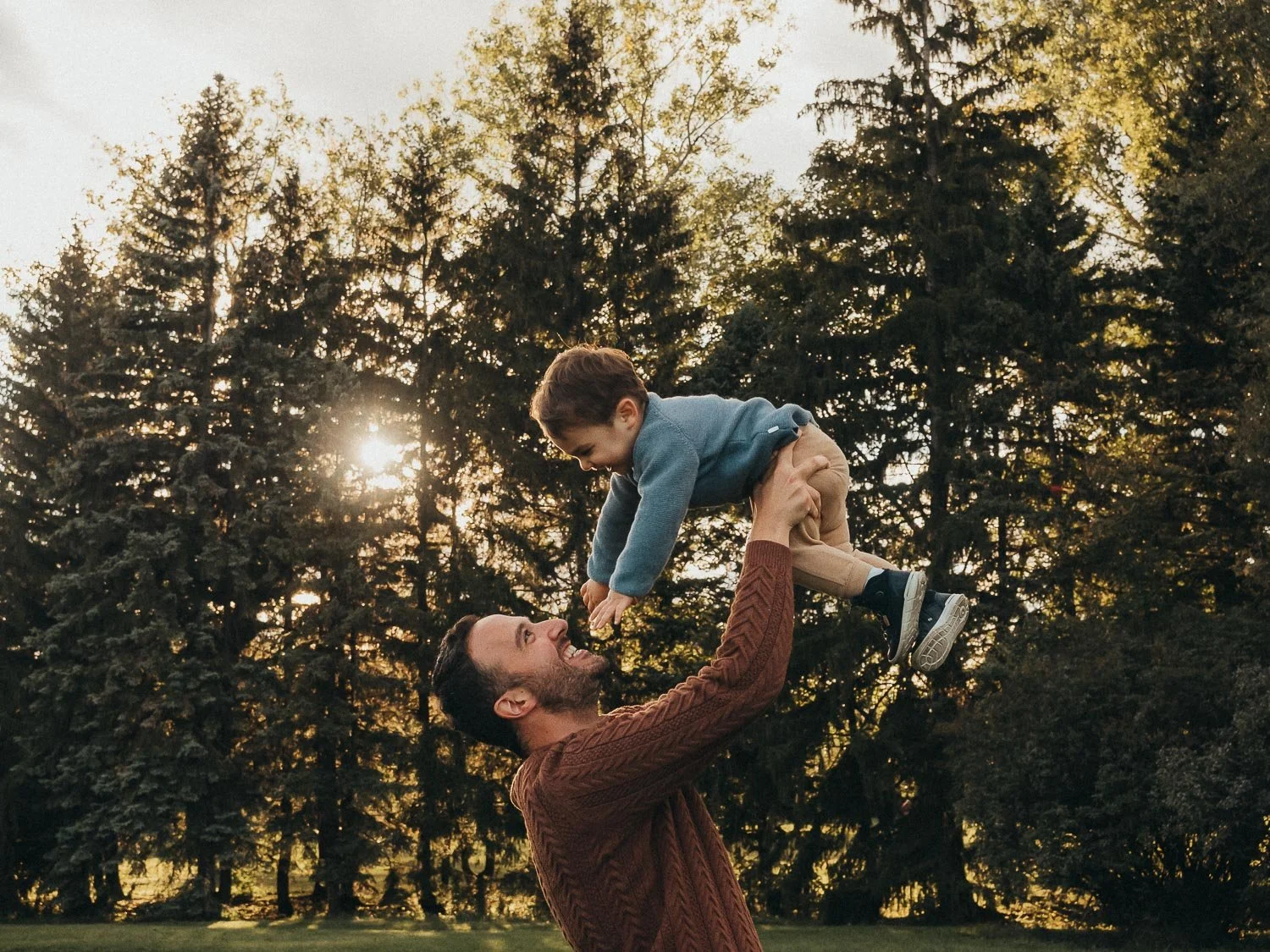 A dad holding his little boy while smiling at him at an outdoor family photoshoot at Maisonneuve Park, Montreal