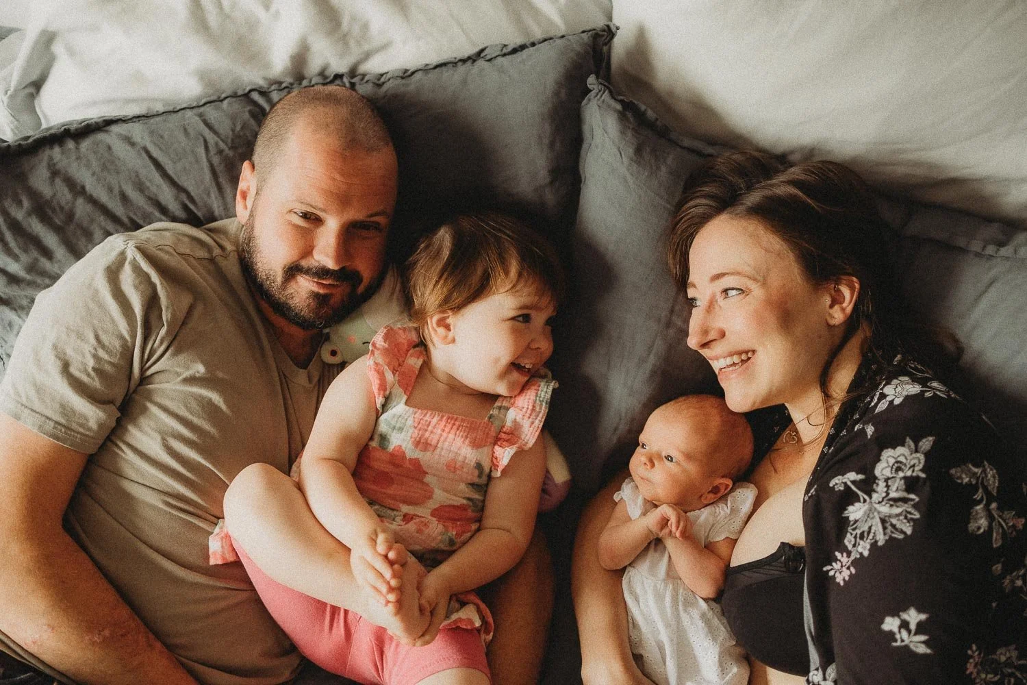 A photo taken from the top of a mom, dad, toddler girl and newborn baby girl, laying down in bes and laughing, captured during an in-home newborn photoshoot in La Salle, Montreal