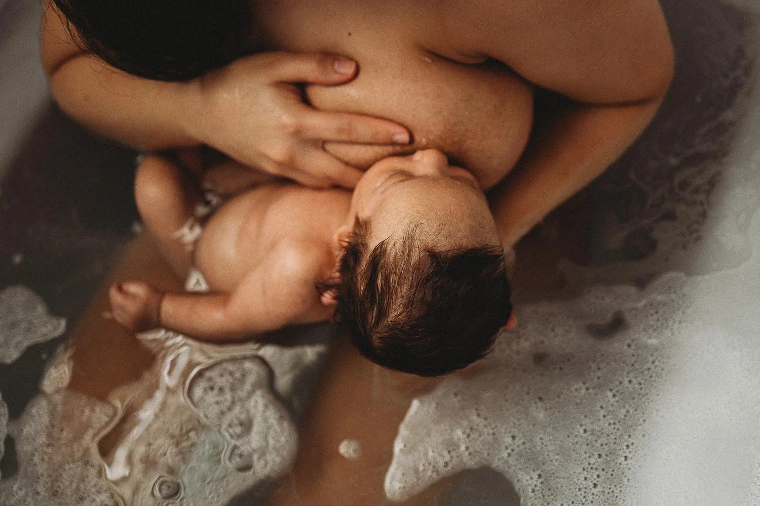 A mom breastfeeding her baby in the bath while holding her breast, captured during an in-home  motherhood photoshoot in St-Constance, Quebec