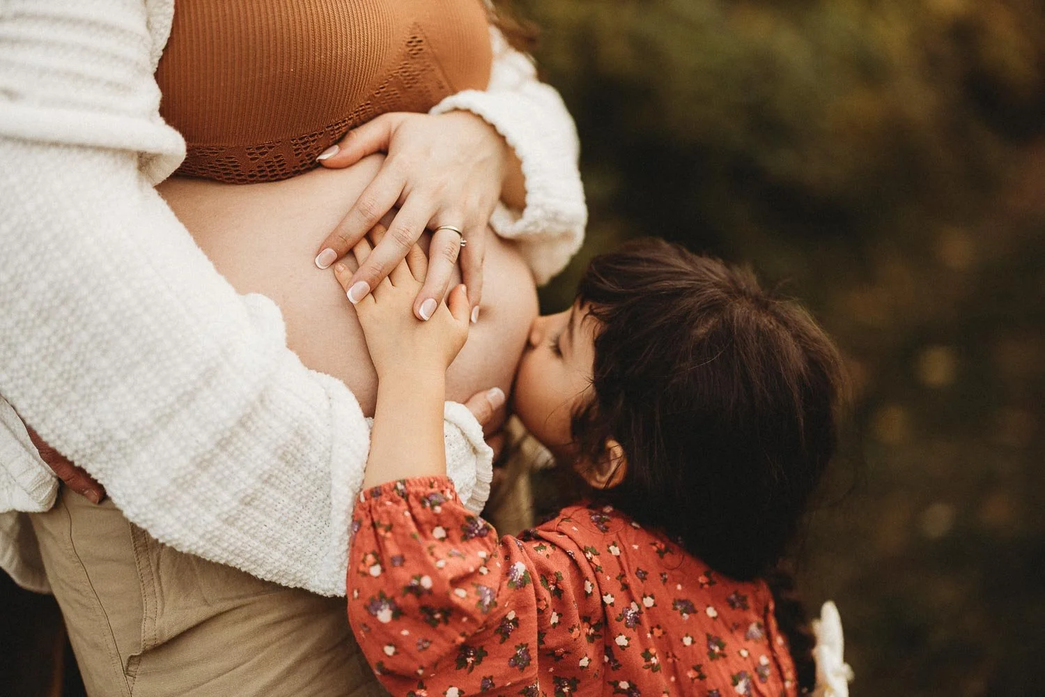 Pregnant mom being kissed in the belly by her young daughter while both holding hands tenderly at an outdoor Maternity Photoshoot at the Boucherville Island, Montreal