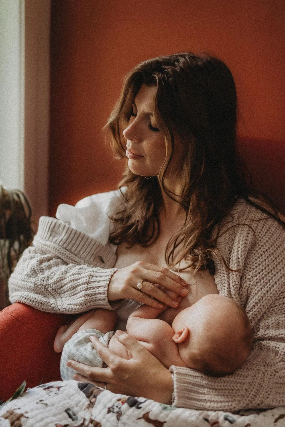 mama breastfeeding her baby while holding his little hand and enjoying it with closed eyes, captured at an in-home newborn photoshoot in Dorval, Montreal