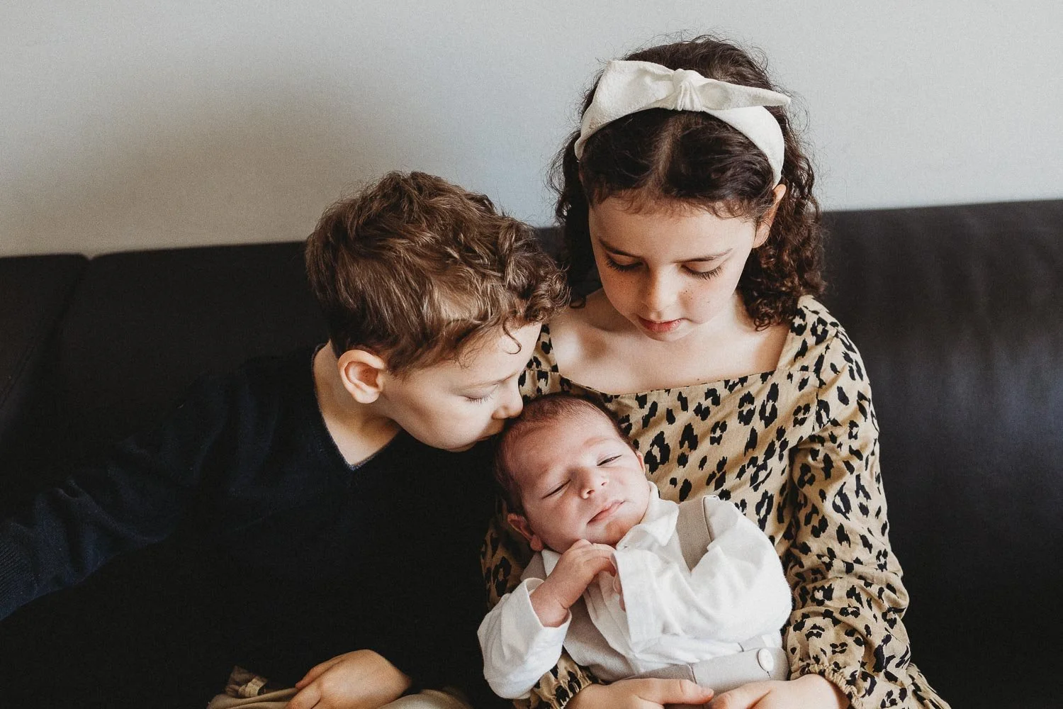 Two children, a girl and a boy, are sitting on a black couch with a newborn baby. The girl is wearing a leopard print top and a white headband, and the boy is wearing a black shirt. The girl is holding the baby and looking down at it, while the boy is leaning in to kiss the baby's head.
