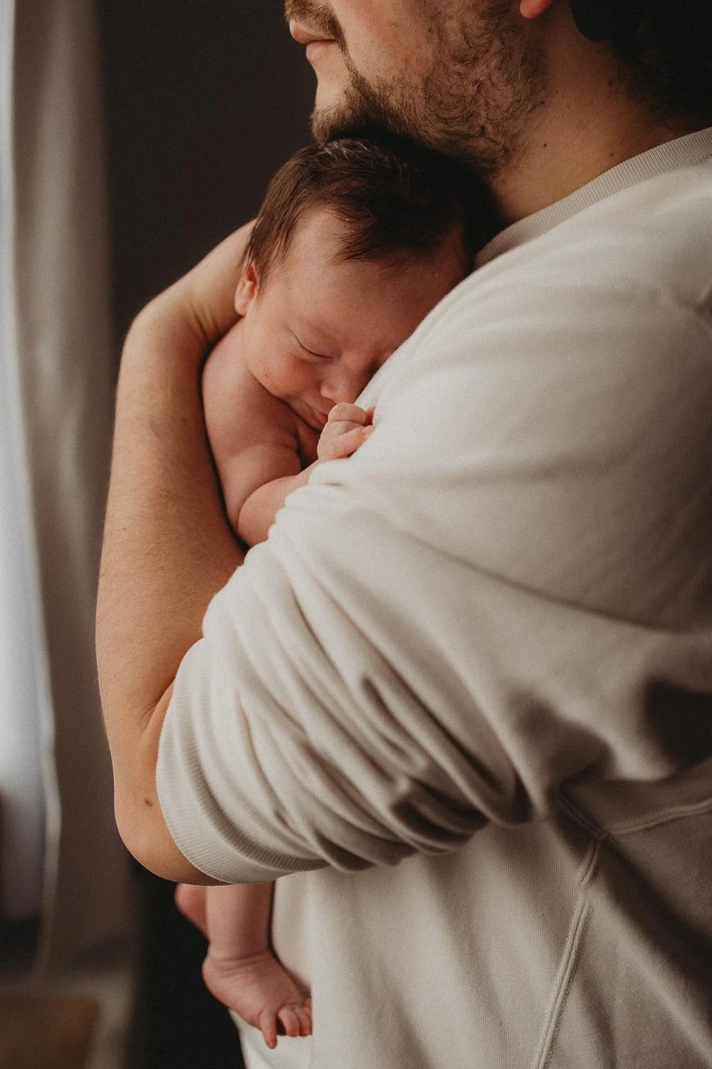 A closed-up photo of a man holding his baby who is resting on his chest, in front of a window during an in-home newborn photoshoot in La Salle, Montreal