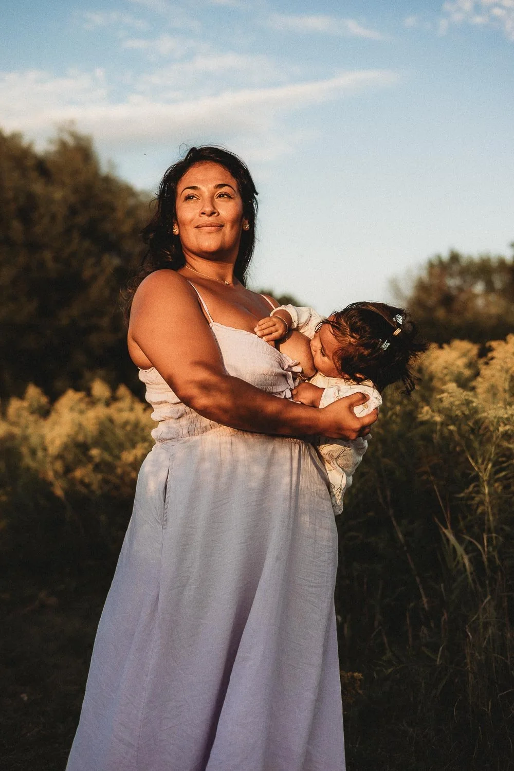 A woman holding a baby girl outdoors during sunset, with trees and a blue sky in the background during a motherhood photoshoot at Boucherville Island