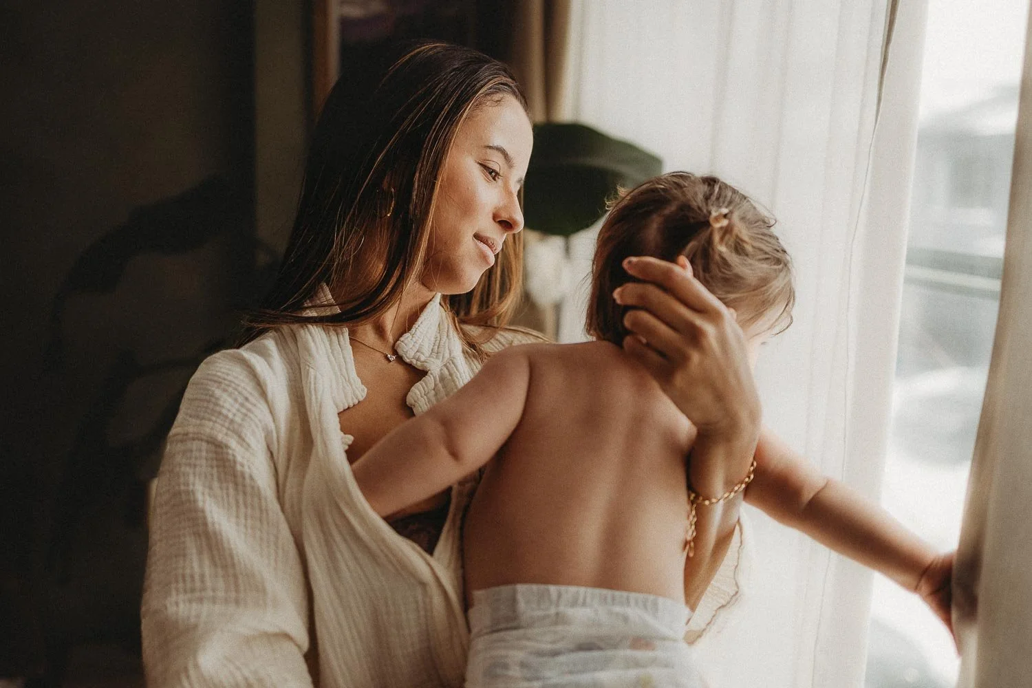 A woman with long brown hair gently holds a young child with curly hair as they look out a window, with warm natural lighting creating a soft, intimate atmosphere captured during an intimate motherhood photo session in Montreal.