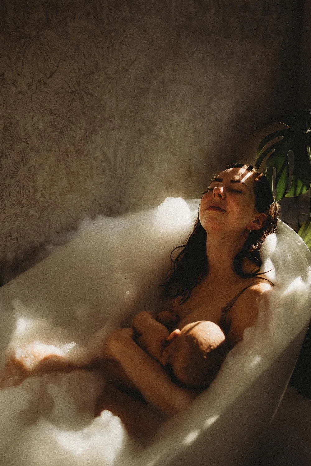 A Dorval motherhood photo of a mom relaxing in a bubble bath with sunlight casting shadows on her face and a plant in the background while breastfeeding her baby
