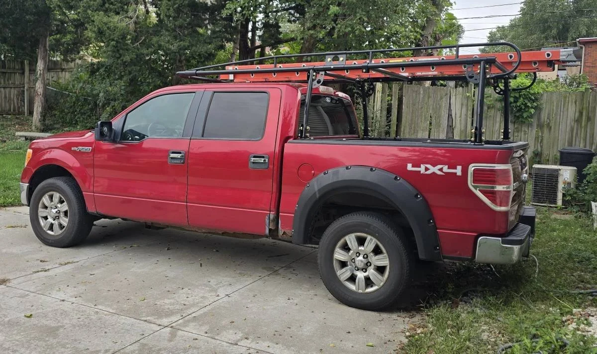 Red F-150 work truck with ladder rack and extension ladder parked in driveway, used by a local roofing and exterior repair contractor in Omaha, Nebraska.