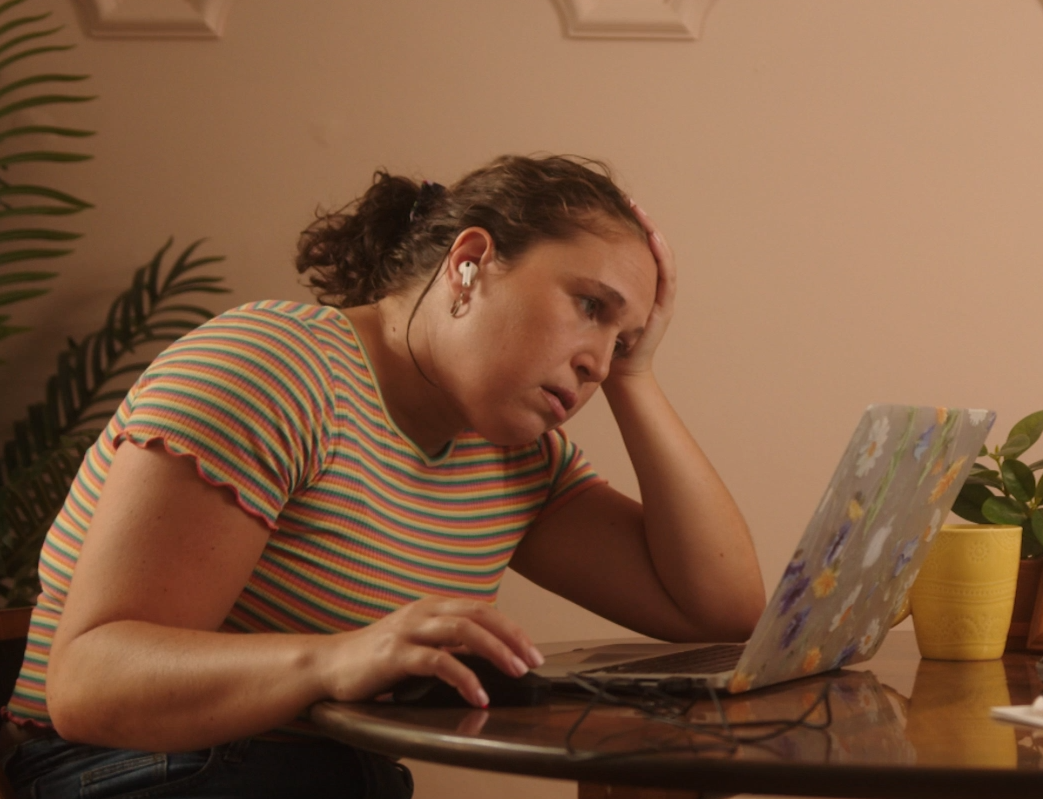 Person in a striped shirt using a laptop, looking frustrated and wearing earbuds.