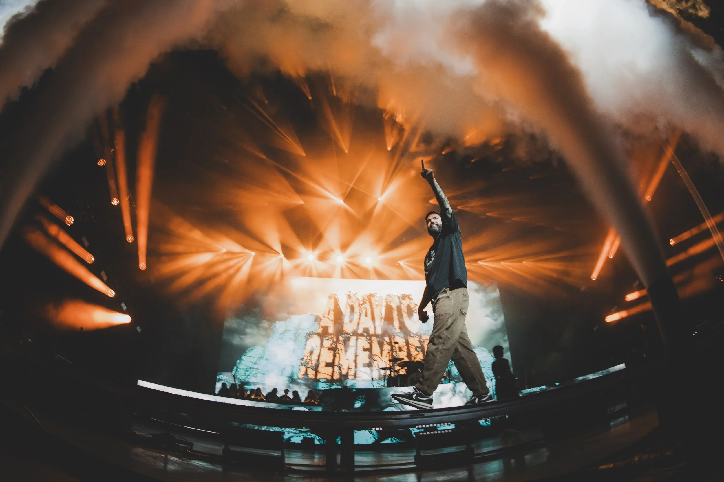 A male performer on stage during a concert, pointing upward with his right hand, with colorful lighting and smoke effects, and a large screen displaying visuals behind him.