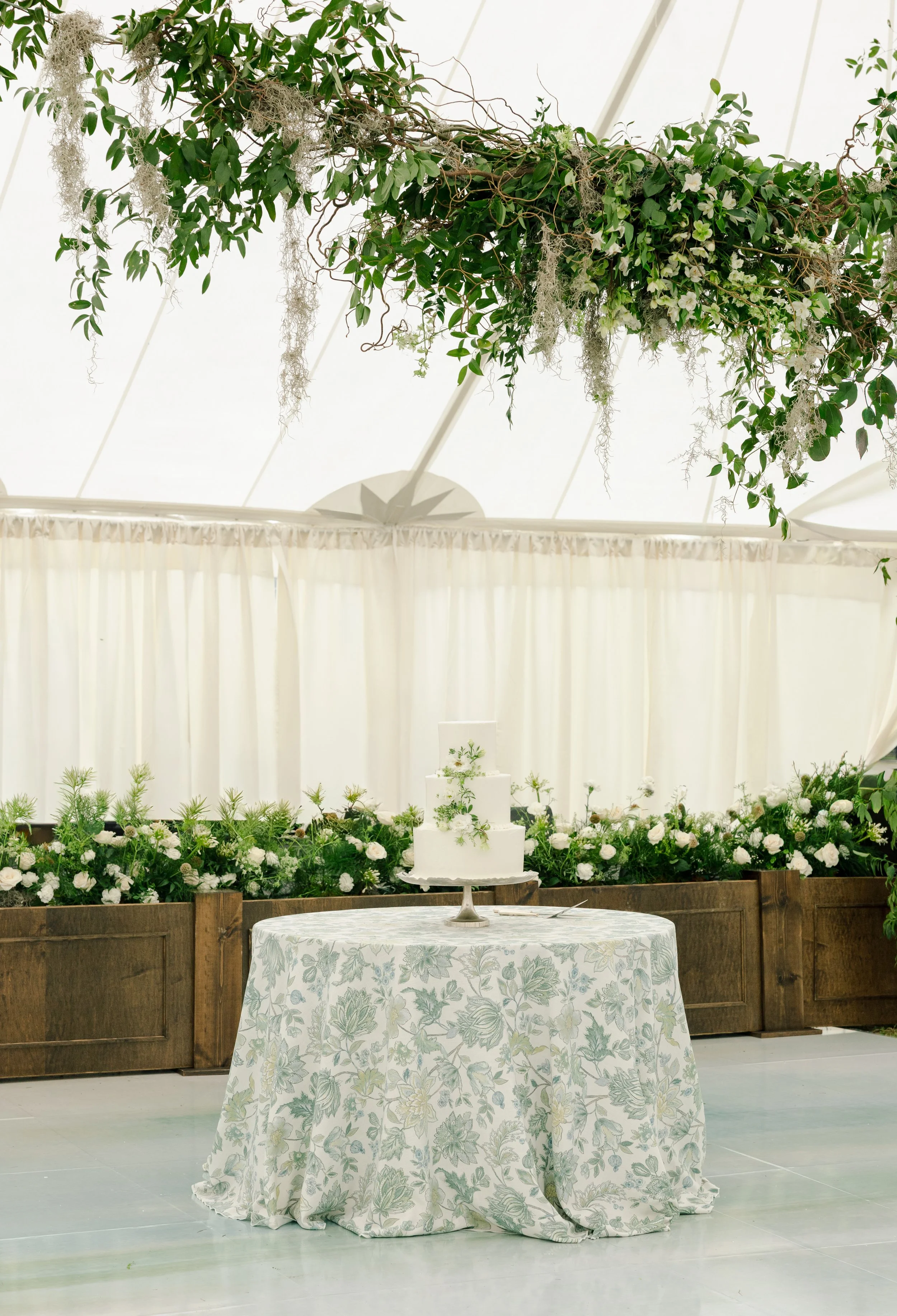 A wedding cake placed on a cake table with a blue and green patterned linen, in the back there is an oak stage facade with florals on top and a band stage set up.