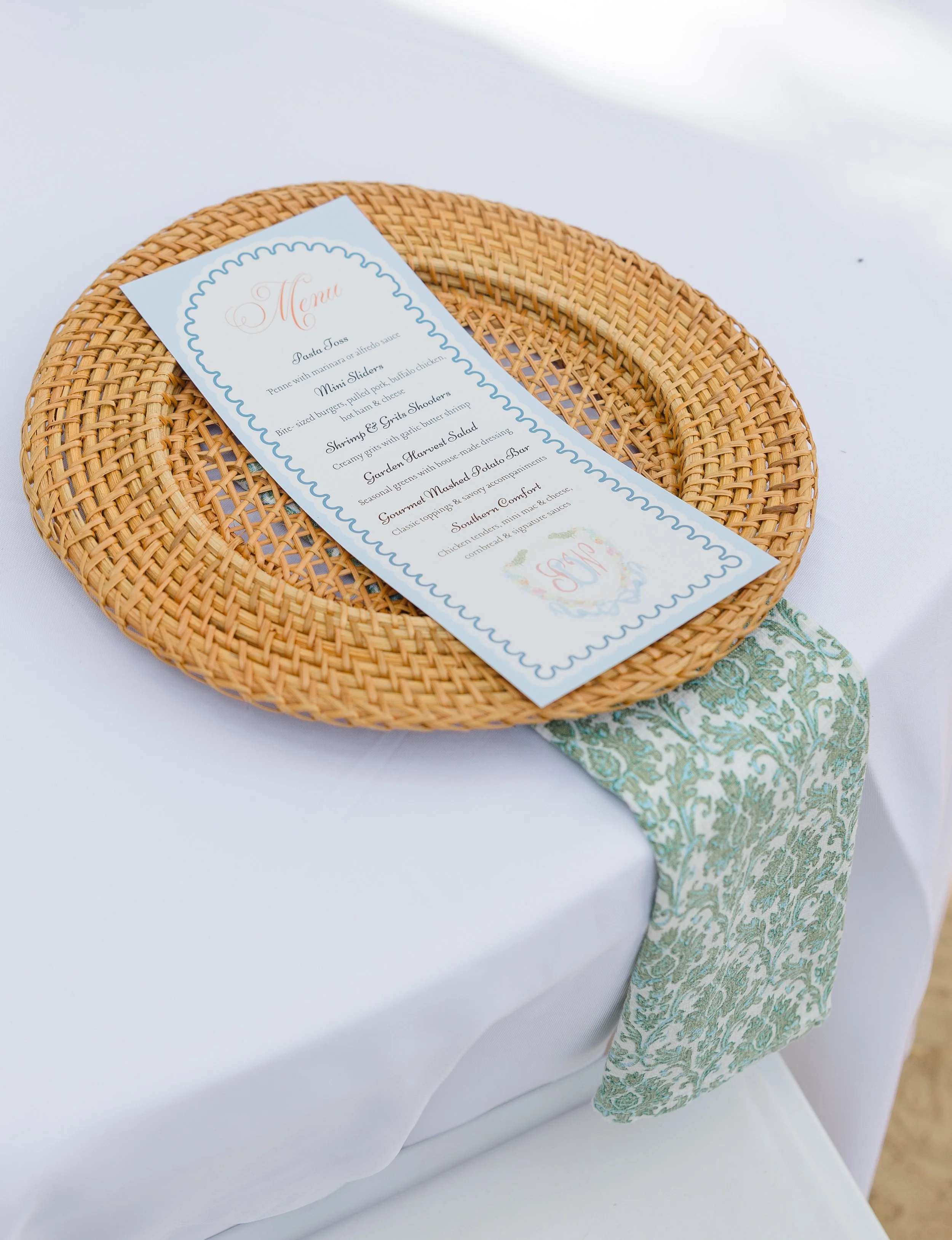 Green and ivory block-print linen napkin draped beneath a woven rattan charger at an elegant wedding place setting.