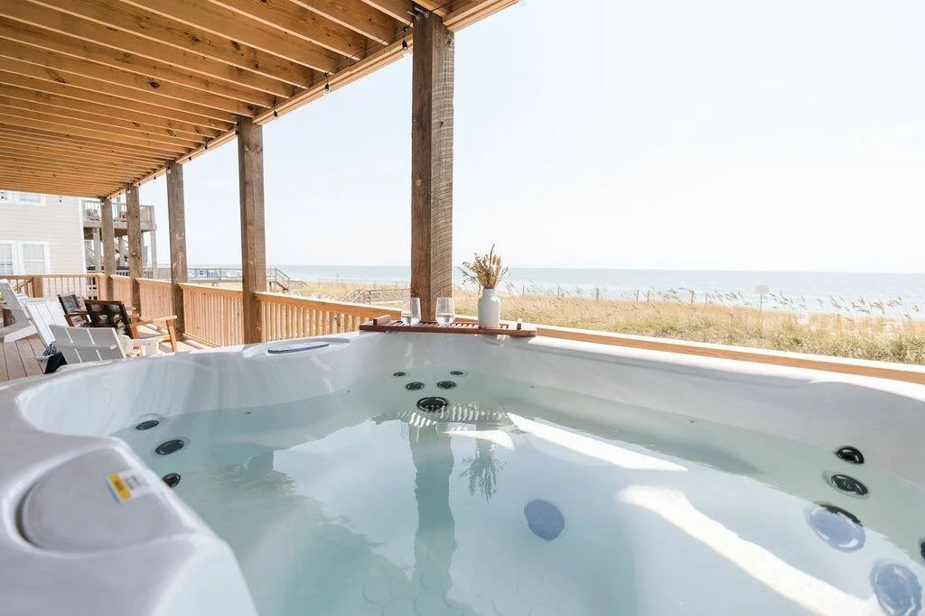Hot tub on a covered porch overlooking a beach and ocean with chairs, a tray with glasses, and a vase with dried flowers.