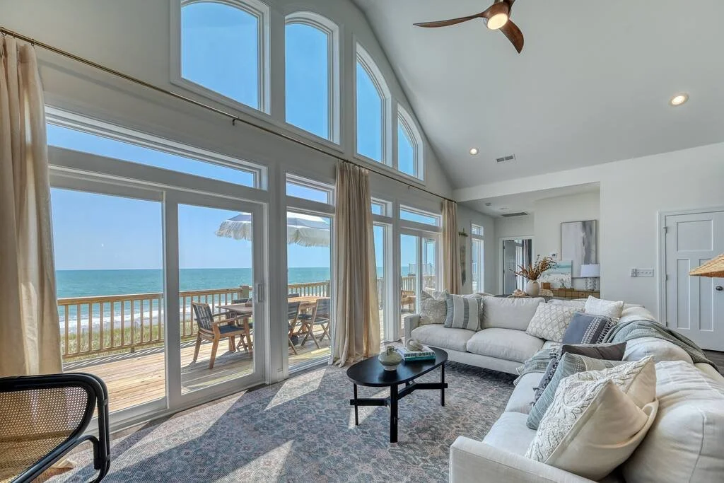 Living room with large windows and sliding glass door overlooking a beach and ocean, with an outdoor patio and dining set, and a white sectional sofa with pillows.