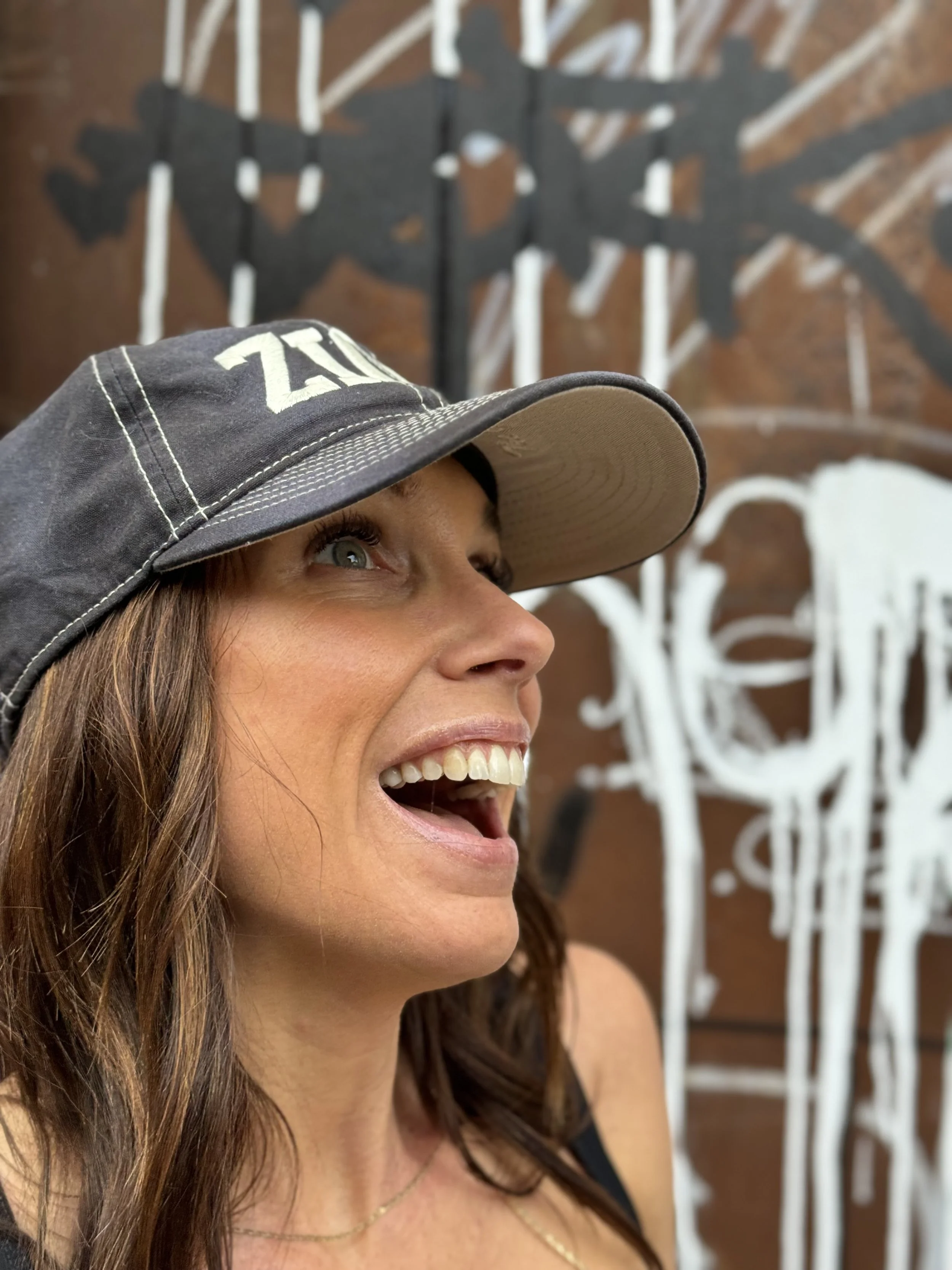 Close-up of a woman with brown hair, wearing a gray cap, smiling and looking upward, with an abstract black and white mural on a wooden wall in the background.