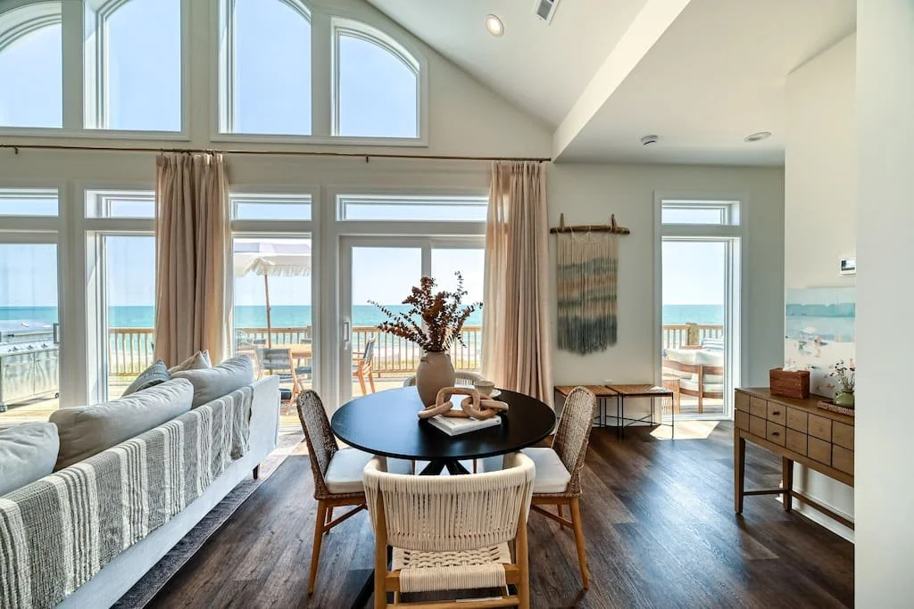 Living room with large windows and ocean view, round dining table with chairs, sofa, and outdoor deck with seating and umbrella.