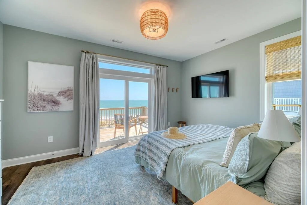 Beachside bedroom with sliding glass door to balcony, ocean view, pastel-colored walls, bed with pillows and blanket, wall-mounted TV, window with bamboo shade, and pendant ceiling light.