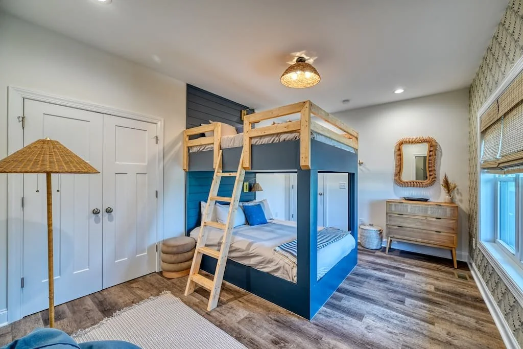 Bedroom with a blue bunk bed, a wooden ladder, a large window with bamboo shades, a mirror on the wall, a wooden dresser, and a standing lamp with a woven shade.