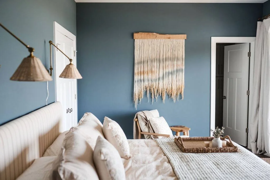 Bedroom with a blue wall, cream-colored bed and pillows, woven wall hanging, white door, window with white curtains, and a wooden chair with blanket and small side table, decorated in neutral and earthy tones.