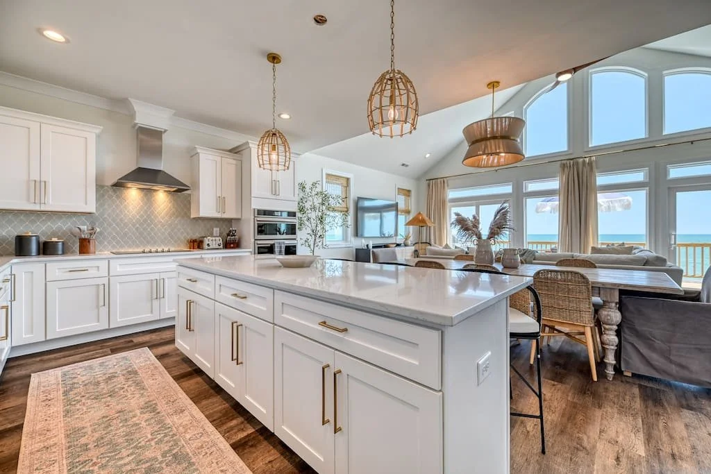 Open-concept kitchen and living area with ocean view, white cabinetry, marble island, pendant lights, and large windows.
