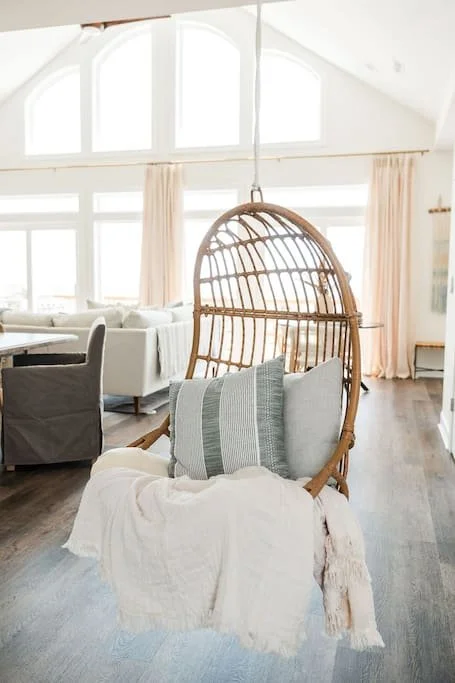A hanging wicker chair with cushions and a blanket in a bright living room with large windows and white curtains.