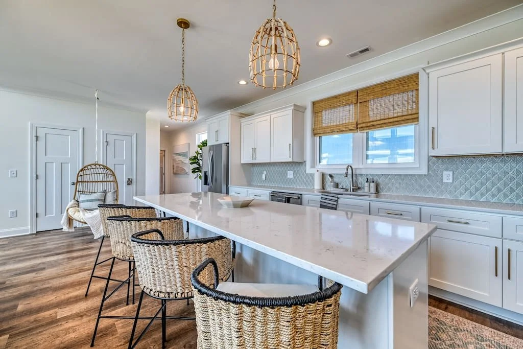 Bright modern kitchen with white cabinets, a large island with a white marble countertop, wicker barstools, pendant lights, and a window with bamboo shades.